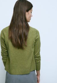 Donna con lunghi capelli castani, che indossa un maglione verde oliva e una gonna grigia, vista di lato su uno sfondo chiaro e semplice.