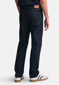 Jeans in denim blu scuro con vestibilità dritta, cuciture in contrasto e una patch in pelle con il logo del marchio sulla vita posteriore. Indossati con scarpe da ginnastica bianche.