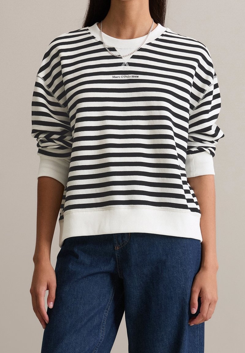 Femme portant un sweat-shirt à rayures horizontales noires et blanches avec le texte Marc O'Polo DENIM et un pantalon en jean bleu foncé, debout les bras détendus.