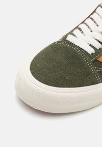 Zapatillas de gamuza verde con suela de goma blanca, que presentan acentos de cuero marrón en contraste y cordones blancos, diseño de punta redondeada.
