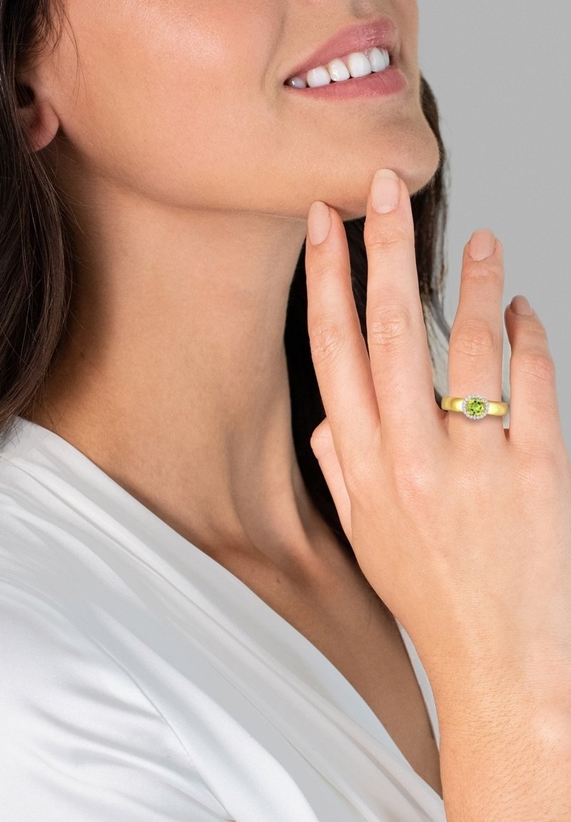 Bague en or jaune avec une pierre verte centrale, entourée de petites pierres claires. La main du modèle est posée sur le menton contre un fond neutre.