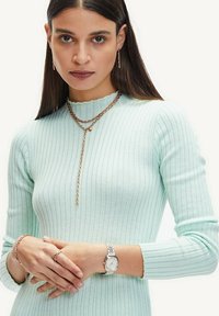 Mintgroene geribbelde gebreide trui met een geschulpte halslijn. Sieraden zijn onder andere lagen rosegouden kettingen, een armband en een zilveren horloge.