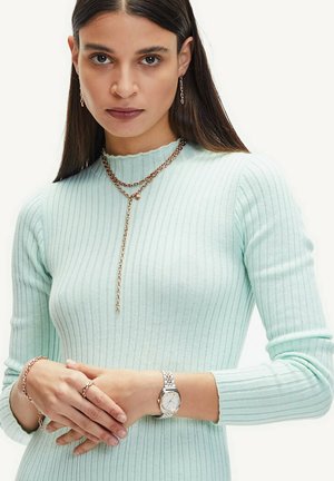 Mintgrüner, gerippter Strickpullover mit einem gewellten Ausschnitt. Die Schmuckstücke umfassen geschichtete Halsketten aus Roségold, ein Armband und eine silberne Uhr.
