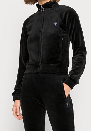 Femme portant une veste zippée en velours noire et un pantalon assorti avec de petits logos bleus brodés, main dans la poche, debout devant un fond blanc.