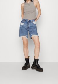 Grå ärmlös topp, högmidjade blå denimshorts med fransade kanter och slitna detaljer, svarta snörstövlar med tjock sula.