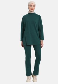 Teal, ribbed turtleneck top with loose fit and long sleeves, paired with matching, fitted pants. Worn with a light beige hijab and beige sneakers.