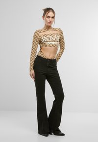 Haut cropped à manches longues beige avec impression de logo, associé à un jean noir évasé ; le mannequin pose devant un fond neutre.