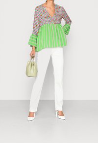 Blouse colorée avec un décolleté en V profond, des manches longues évasées et un mélange de motifs roses, rouges et verts ; associée à un pantalon blanc et un sac vert clair.