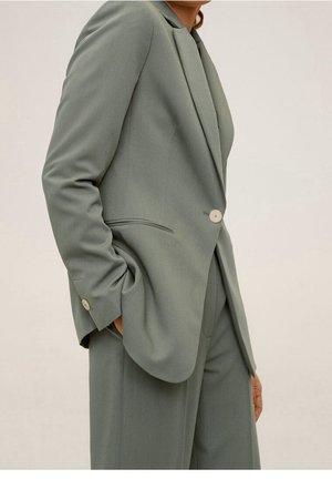 Personne portant un costume gris-vert clair sur mesure avec un blazer à un seul bouton et un pantalon large assorti, main dans la poche du blazer.