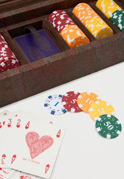 Jetons de poker de différentes couleurs et valeurs, un étui à jetons à motif cachemire, et un éventail de cartes à jouer avec des cœurs sur une surface blanche.