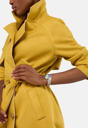Yellow ribbed coat with a high collar, cinched waist, front buttons, and two pockets. Silver watch with a multicolored dial on wrist.