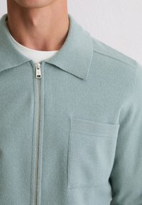 Jasnoniebieska, rozpinana kurtka wykonana z miękkiego, teksturowanego materiału. Posiada pointed kołnierz oraz kieszeń na piersi. Srebrny zamek błyskawiczny jako akcent.