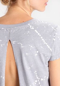 Šedé tričko s designem výřezem na zádech. Obsahuje bílé skvrny rozptýlené po látce. Měkký materiál s krátkými rukávy.