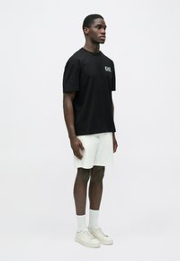 Férfi áll előre nézve, fekete EA7 pólót, fehér rövidnadrágot, fehér zoknit és fehér tornacipőt visel, sima világos háttér előtt.