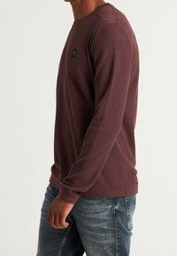 Chemise à manches longues en tricot gaufré couleur bordeaux, dotée d'un col rond et d'un petit écusson logo noir sur la poitrine gauche. Texture douce.