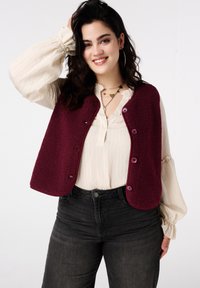 Veste sans manches en polaire bordeaux à la texture douce, avec des boutons violets et des bords arrondis. Portée sur un chemisier crème à motif plissé.
