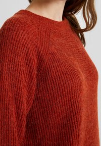 Pull tricoté couleur rouille avec texture côtelée et encolure arrondie, présentant des points visibles qui soulignent son matériau doux et duveteux.