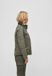 Olive green utility jacket with a zip front, snap pockets, and a high collar, featuring a textured fabric and structured silhouette.