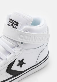 Zapatillas altas blancas de piel sintética suave, con un logotipo de estrella negra, costuras en contraste y una correa de velcro.