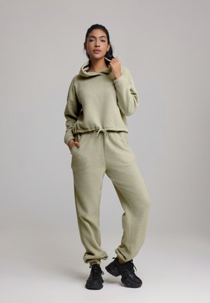 Ensemble de loungewear en polaire kaki avec capuche, comprenant un haut court et un pantalon à taille élastique. Porté avec des chaussures de sport noires.