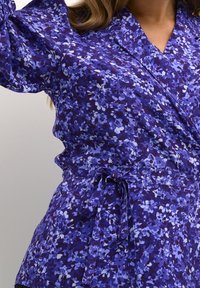 Haut cache-cœur à fleurs violettes en tissu texturé avec des nuances de bleu et de blanc. Comprend une ceinture à la taille et un décolleté en V.