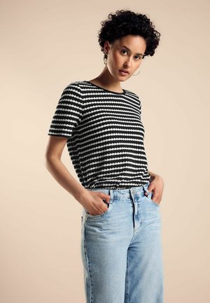 Gestreifte schwarz-weiße Kurzarm-T-Shirt mit rundem Halsausschnitt, kombiniert mit hellblauen Jeans. Die Hände des Modells sind in den Taschen.
