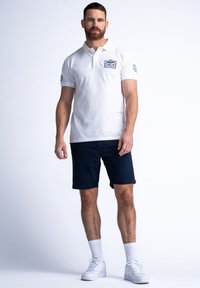 Bílé polo tričko s krátkým rukávem, s modrou nášivkou na prsou, v kombinaci s námořnickými šortkami a bílými teniskami.