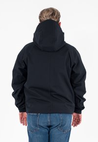 Persoon met kort haar, gekleed in een zwart met capuchon jack en blauwe jeans, gezien van achteren tegen een witte achtergrond.