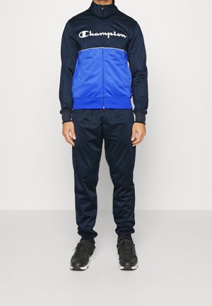 Mann trägt eine marineblaue und blaue Champion-Trainingsjacke mit Reißverschluss, dunkelblaue Trainingshose und schwarze Turnschuhe, steht vor einem schlichten Hintergrund.