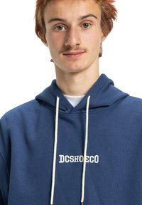 Námornícky modrá mikina s kapucňou z bavlny, s predným logom "DCSHOECO" v bielom vyšívaní, s kapucňou na šnúrku a jemnou textúrou.