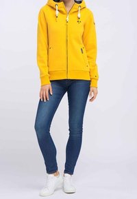 Felpa gialla in pile con zip, due tasche frontali, coulisse regolabili, abbinata a jeans skinny blu e sneakers bianche.