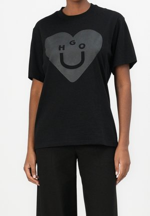 Person trägt ein schwarzes T-Shirt mit großem grauem Herz und dem Schriftzug "HUGO", der auf der Vorderseite ein Lächeln bildet, kombiniert mit schwarzer Hose.