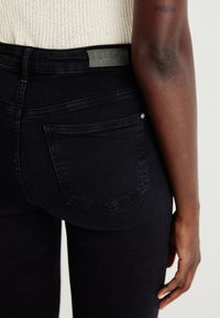 Skinny black jeans featuring a high waistband, back pockets, and a branded label. The fabric has a smooth, slightly stretchy texture.