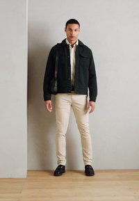 Veste texturée vert foncé sur une chemise blanche, pantalon beige ajusté et chaussures noires. Le mannequin tient un sac noir et se tient sur un sol en bois.