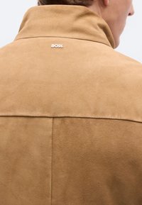 Rjavi antilop usnjen jopič z visokim ovratnikom, ki ima podrobnosti šivanja in majhen logotip "BOSS" na hrbtni strani. Mehka tekstura, minimalističen dizajn.