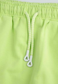 Jasnozielone szorty z elastycznym paskiem w talii. Zawierają białe plecione sznurki ze plastikowymi końcówkami. Miękka, gładka tekstura materiału.
