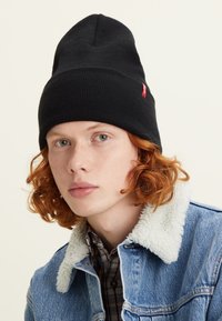 Black knit beanie with a tapered top and a small red brand label. Worn by a person with curly red hair, layered under a denim jacket.