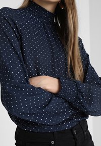 Mörkblå blus med vita prickar, med en smockad hög hals och volangärmar, tillverkad av ett slätt tyg.