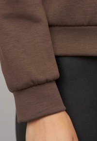 Brauner Langarmoberteil mit elastischen Bündchen. Das Material wirkt glatt und eng anliegend, kombiniert mit schwarzem Stoff im Hintergrund.
