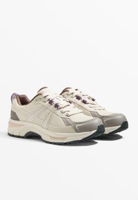 Béžové tenisky s sieťovinou, hnedými akcentmi a fialovými šnúrkami. Hustá, textúrovaná medzipodrážka a čierna gumová podrážka pre trakciu.