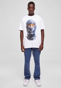 Balta oversized t-kreklu ar zilu grafiku, kurā attēlots maskēts tēls. Kombinēts ar ziliem džinsiem un baltām sporta apaviem.