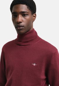 Maglione a collo alto color bordeaux con colletto a coste, texture lavorata a maglia e piccolo logo ricamato sul petto. Design aderente.