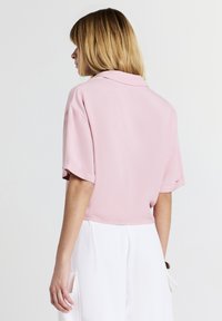 Donna con capelli biondi che indossa una maglietta rosa a maniche corte e larghe e pantaloni bianchi a vita alta, in piedi davanti a uno sfondo bianco uniforme.