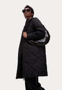 Cappotto lungo nero trapuntato con colletto alto, dotato di una zip a due vie e tasche laterali, abbinato a una borsa a spalla nera lucida.