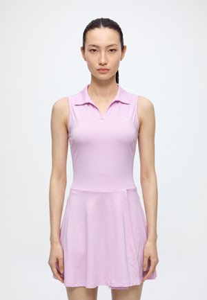 Une femme se tient face à l’objectif devant un fond clair uni. Elle porte un haut rose sans manches à col et une jupe de tennis assortie.