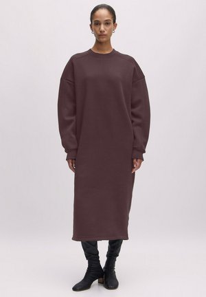 Lange, oversized jurk in een diepe bordeauxkleur, gemaakt van zachte stof met een ronde halslijn en naar beneden gevallen schoudernaden. Subtiele textuur en manchetmouwen.
