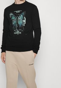 Maglione nero con stampa grafica di ali di farfalla in turchese e blu, con effetti di schizzi aggiuntivi e dettaglio di codice a barre nella parte inferiore.