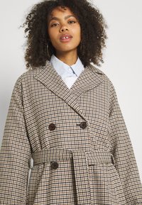 Dvojradový kabát v houndstooth vzore, s opaskom v páse, širokým golierom a hnedými gombíkmi, z texturovanej látky v béžovej a hnedej farbe.