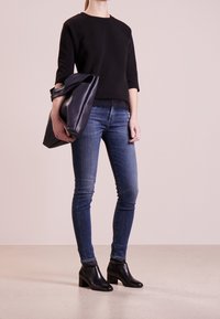 Svart långärmad sweatshirt med spetskanter, parat med blå skinny jeans. Svarta ankelboots med blockklack. Modell som håller en marinblå tote-väska.