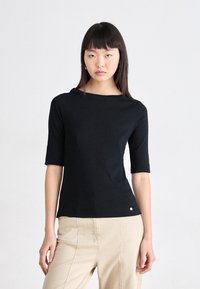 Svart kortermet topp med båthals, myk tekstur og tettsittende design. Har en liten logo ved kanten. Kombinert med lyse beige bukser.